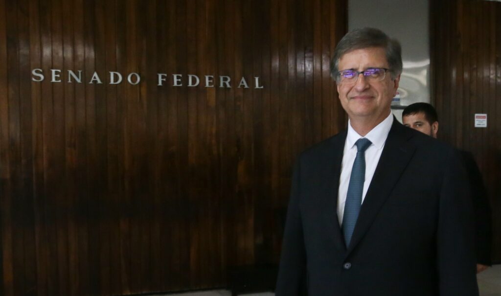 Paulo Gonet (procurador-geral da República) sorrindo em frente a uma parede de madeira com a inscrição "SENADO FEDERAL".