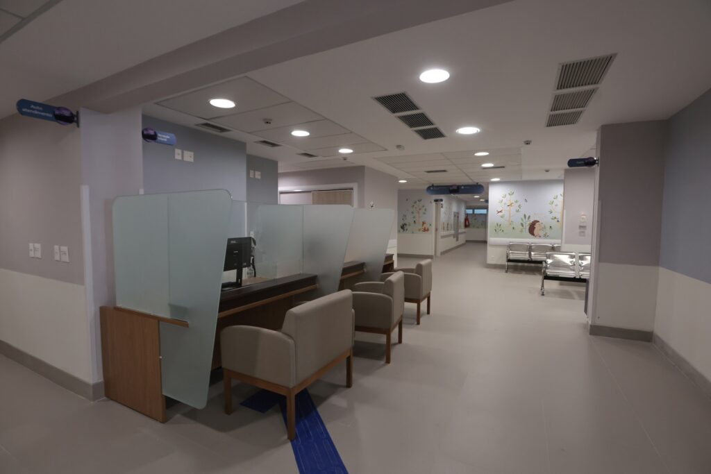 Interior de recepção de hospital com balcões de atendimento, cadeiras bege e murais com desenhos de animais ao fundo.