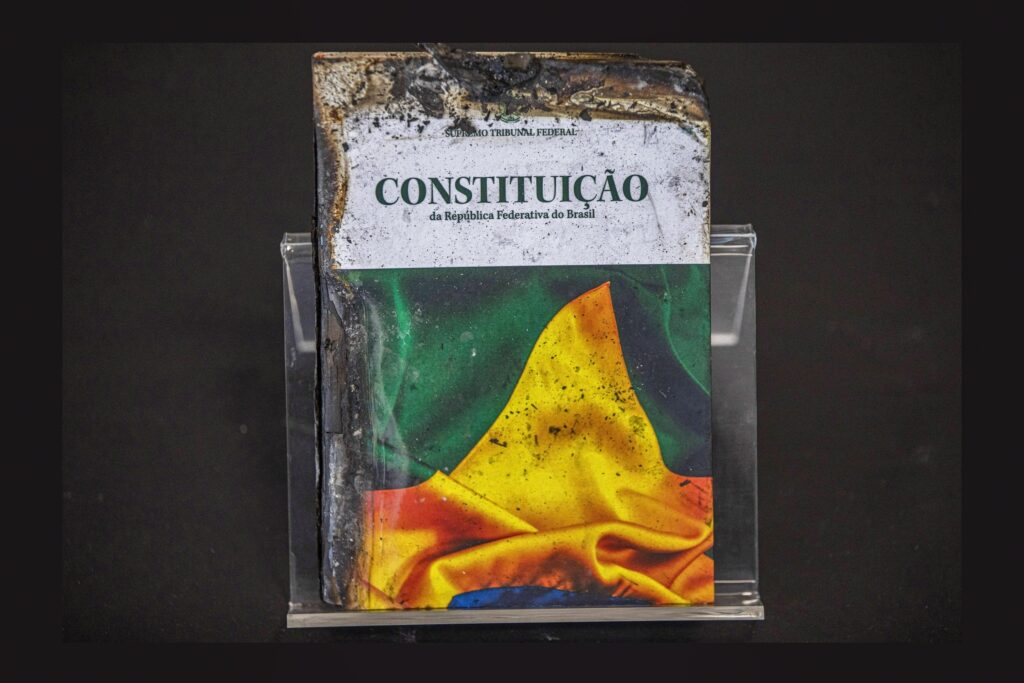 Exemplar carbonizado da 'CONSTITUIÇÃO da República Federativa do Brasil' exposto em suporte de acrílico sobre fundo escuro.