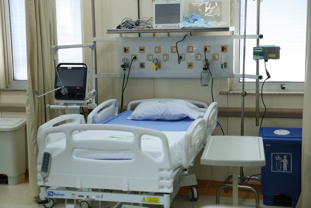 Leito hospitalar com lençol azul e travesseiro branco, cercado por monitores e equipamentos médicos em um quarto de hospital.