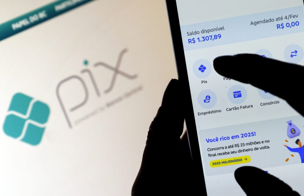Mãos operam aplicativo bancário em smartphone, com o dedo tocando a opção "Pix". Ao fundo, tela de computador com o logotipo do Pix.