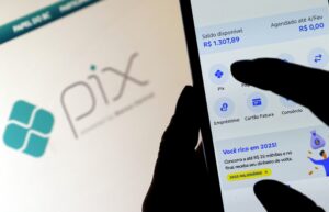 Mãos operam aplicativo bancário em smartphone, com o dedo tocando a opção "Pix". Ao fundo, tela de computador com o logotipo do Pix.