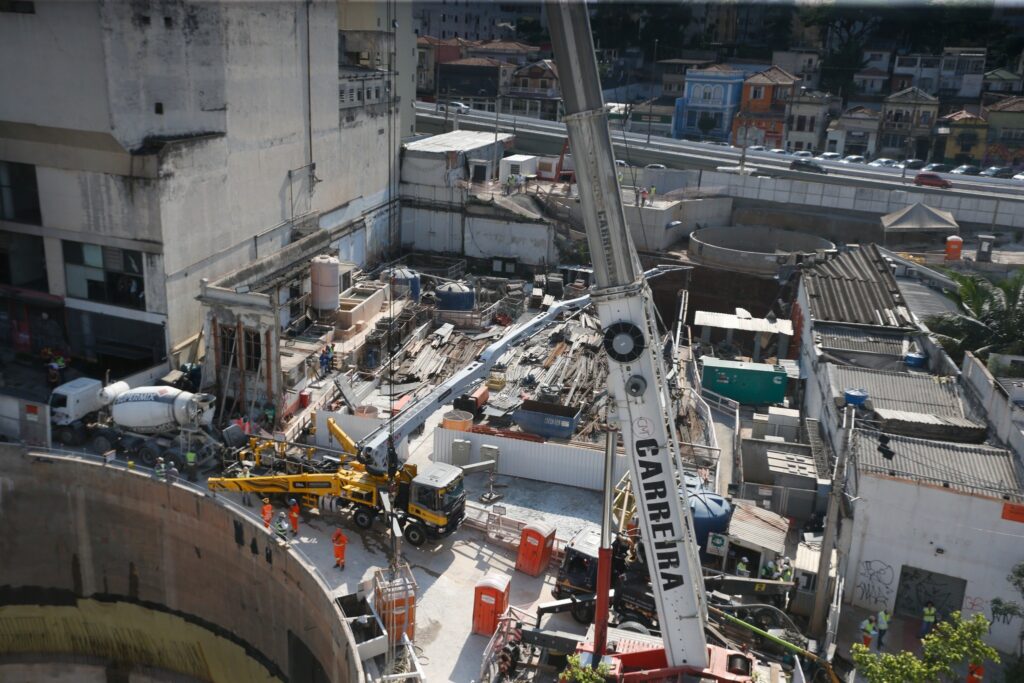 Vista aérea de canteiro de obras com guindastes, caminhão betoneira e operários em área de escavação urbana.