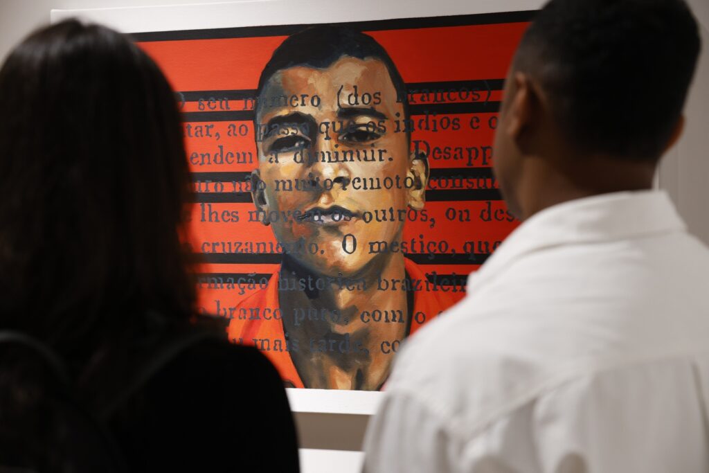 Duas pessoas observam a obra de arte com o rosto de um homem e textos sobre um fundo vermelho, na exposição "Encruzilhadas da Arte Afro-Brasileira".