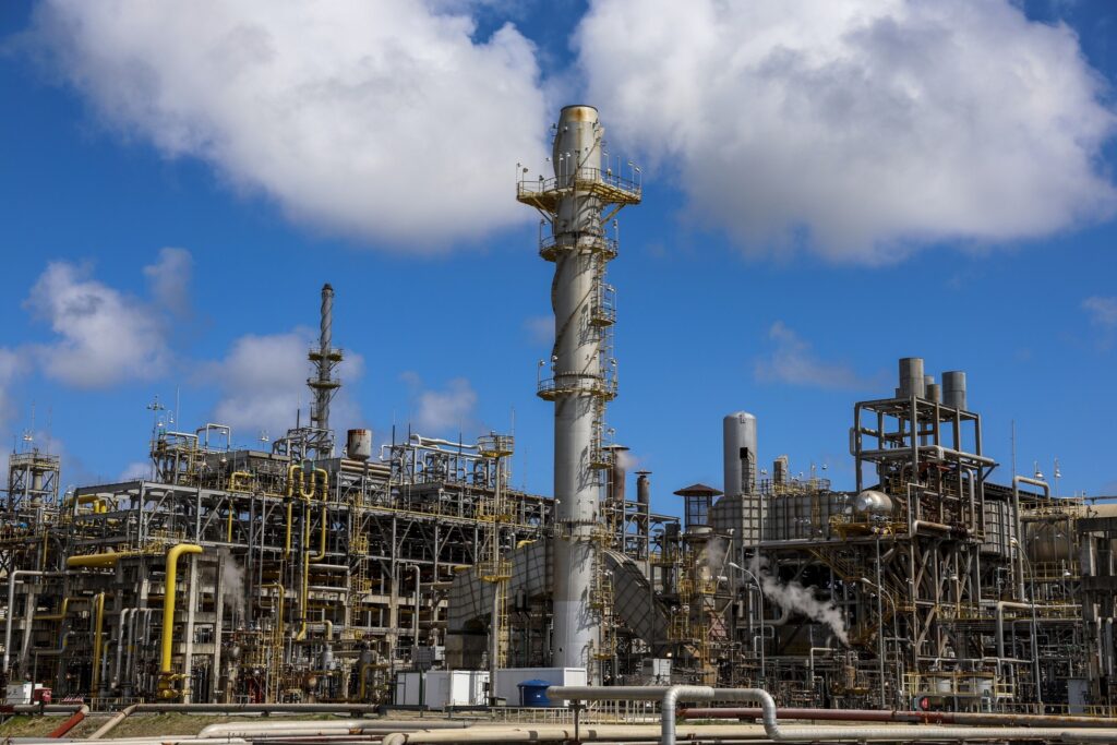 Instalações industriais de uma refinaria com torres altas e rede de tubulações sob um céu azul com nuvens.