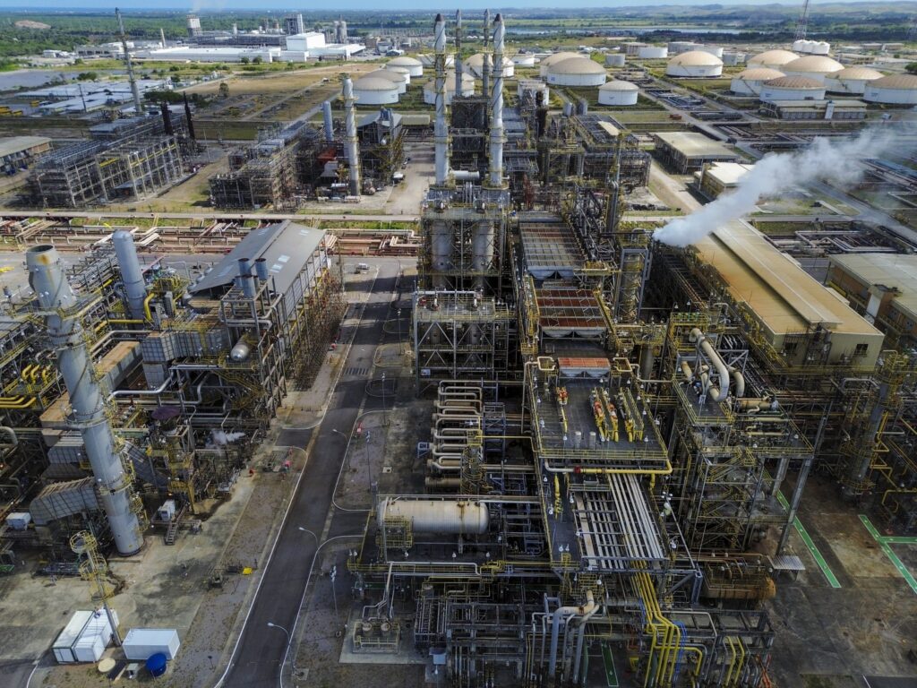 Vista aérea da Refinaria Abreu e Lima, com complexos de tubulações, torres industriais e tanques de armazenamento ao fundo.