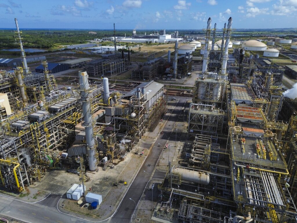 Vista aérea de uma refinaria de petróleo com complexos de tubulações, torres industriais e tanques de armazenamento sob céu azul.