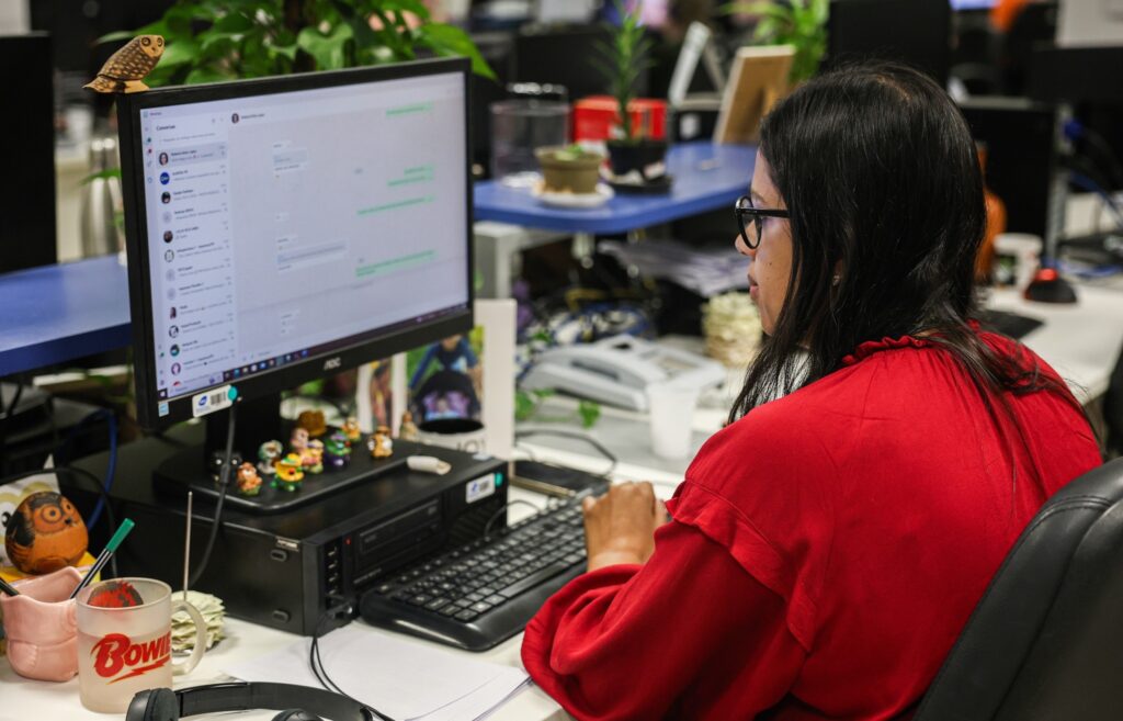 Mulher de blusa vermelha e óculos trabalha em frente a um computador em uma redação.