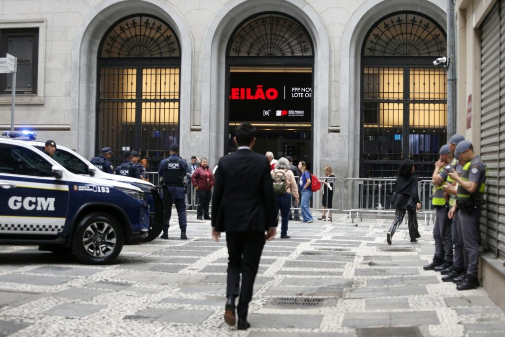 Agentes da GCM e civis em frente ao prédio da B3, com um painel digital exibindo "LEILÃO PPP NOV LOTE OE".