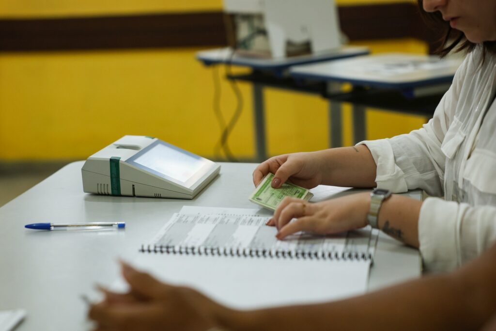 Mãos de uma pessoa segurando um título de eleitor e apontando para uma lista de nomes sobre uma mesa com uma urna eletrônica ao lado.