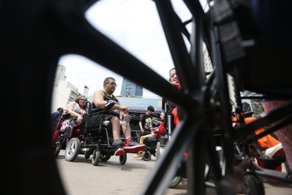 Pessoas em cadeiras de rodas motorizadas reunidas em evento, vistas através dos raios de uma roda, com faixa onde se lê "DO ORGULHO DA DEFICIÊNCIA DE SÃO PAULO".