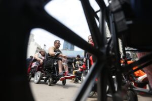 Pessoas em cadeiras de rodas motorizadas reunidas em evento, vistas através dos raios de uma roda, com faixa onde se lê "DO ORGULHO DA DEFICIÊNCIA DE SÃO PAULO".