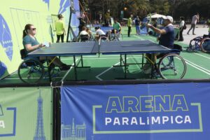 Homens e mulheres em cadeiras de rodas praticam esportes em uma quadra verde; em primeiro plano, uma faixa azul com os dizeres "ARENA PARALÍMPICA".
