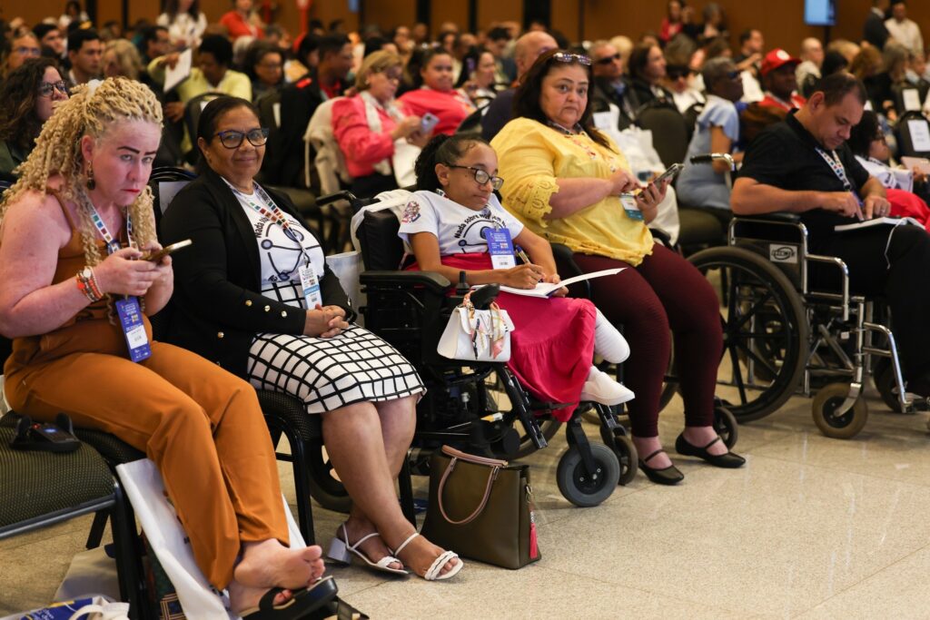 Pessoas com e sem deficiência assistem a uma conferência, com algumas delas em cadeiras de rodas e vestindo camisetas com a frase "Nada Sobre Nós Sem Nós".