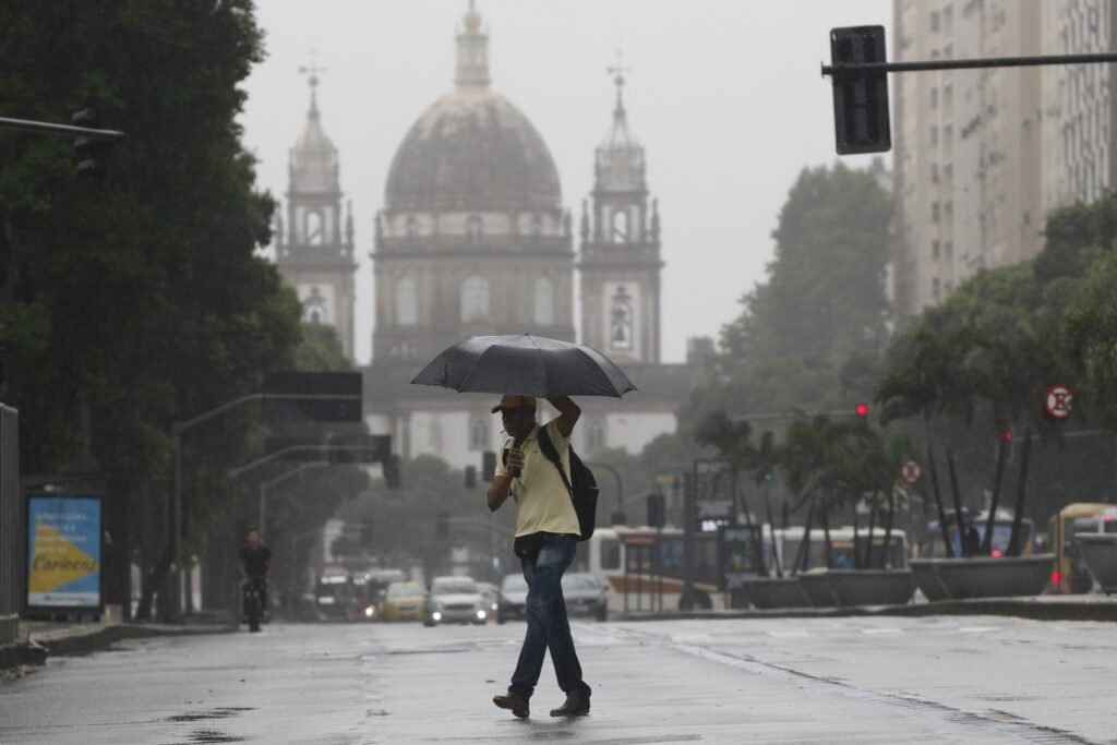 Homem caminha sob guarda-chuva preto em rua molhada com a silhueta de uma catedral ao fundo durante temporal.