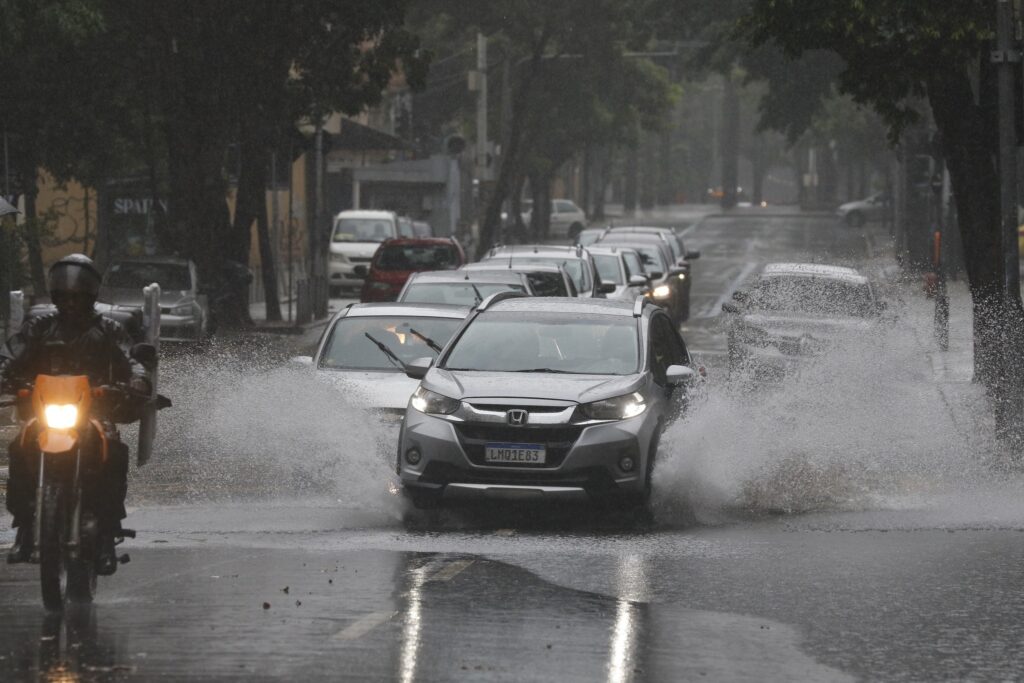 Um carro prata atravessa uma poça de água em uma rua inundada durante forte chuva, provocando respingos, enquanto um motociclista circula ao lado.