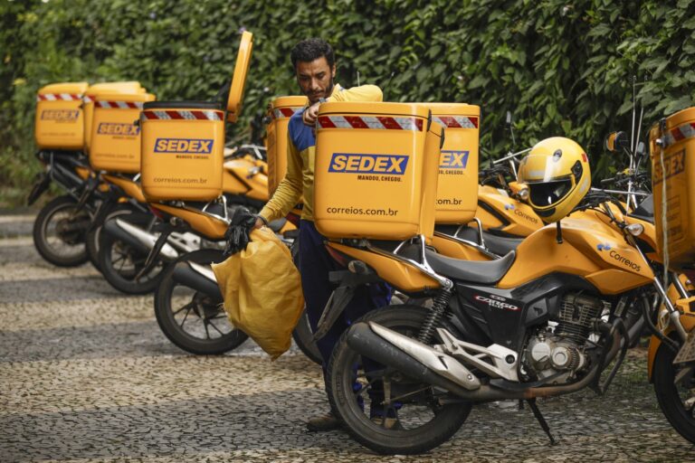 Entregador dos Correios em uniforme amarelo e azul segura uma bolsa amarela ao lado de motocicletas de entrega com a marca "SEDEX".