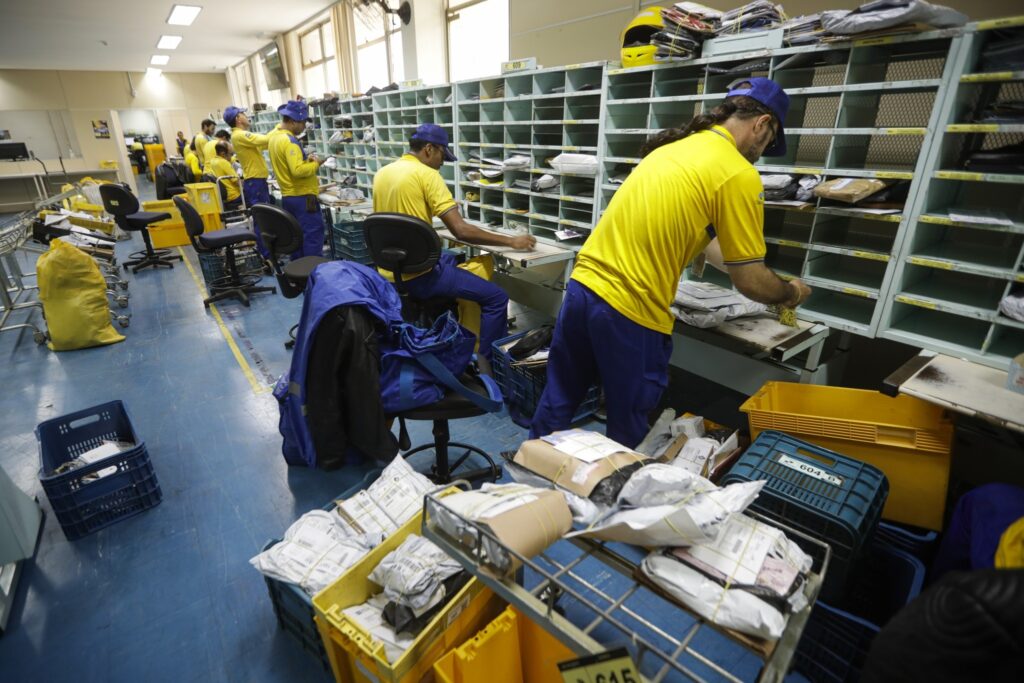 Trabalhadores dos Correios, vestindo uniformes amarelos e azuis, organizam pacotes e correspondências em prateleiras de uma central de distribuição.