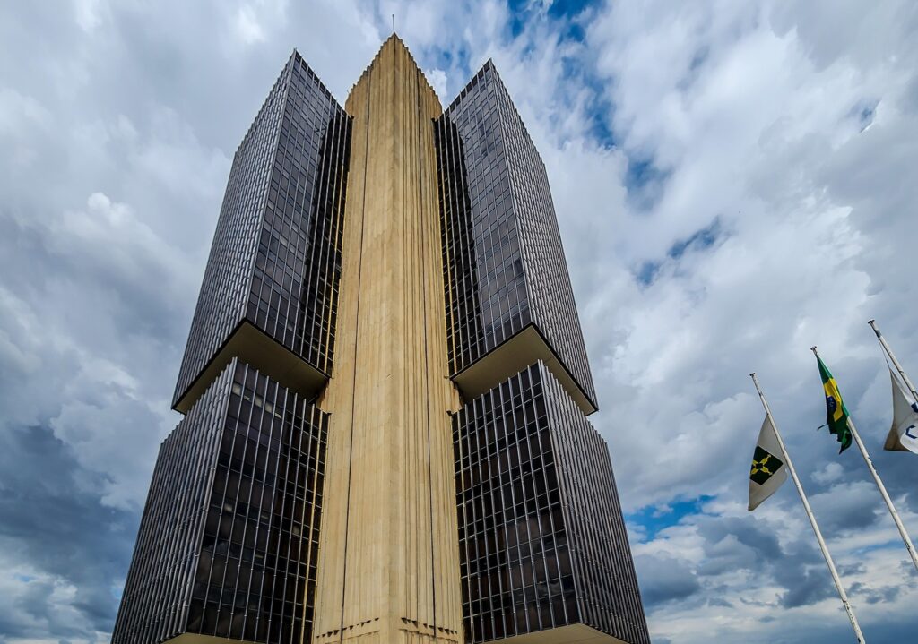 Prédio do Banco Central em Brasília visto de baixo, sob um céu nublado, com a bandeira do Brasil e outras hasteadas ao lado.