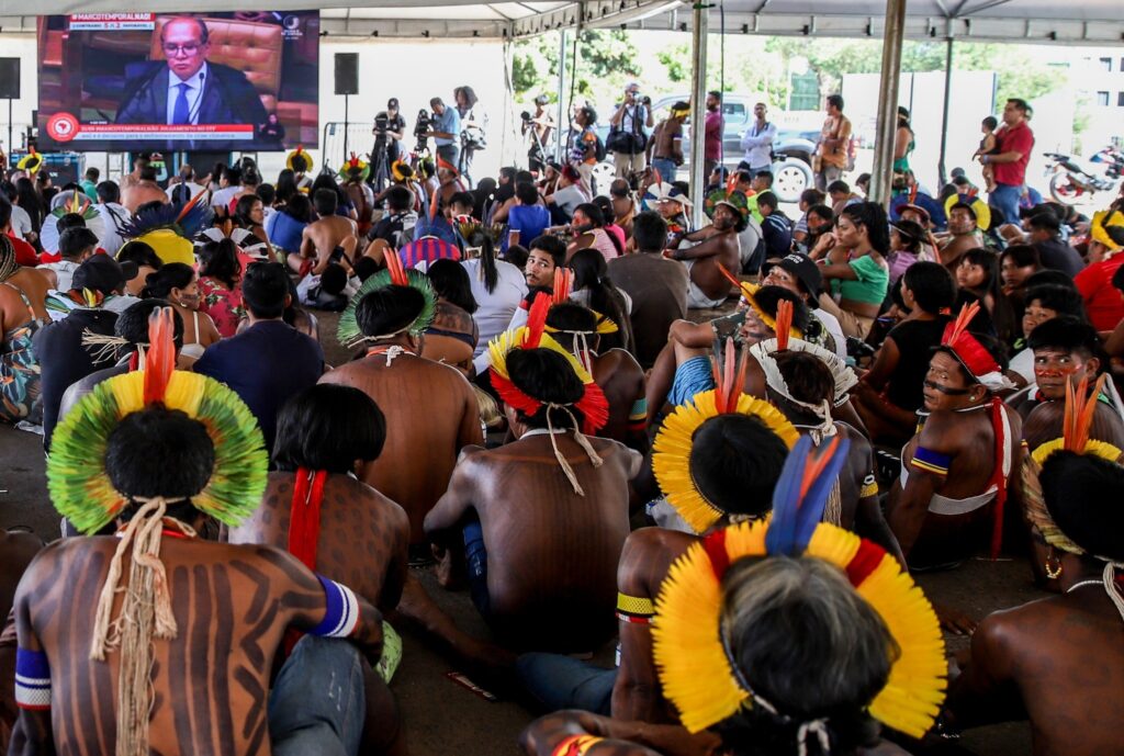 Indígenas com cocares e pinturas corporais sentados sob uma tenda assistem a uma transmissão em telão com os dizeres "#MARCOTEMPORALNAO" e "JULGAMENTO NO STF".