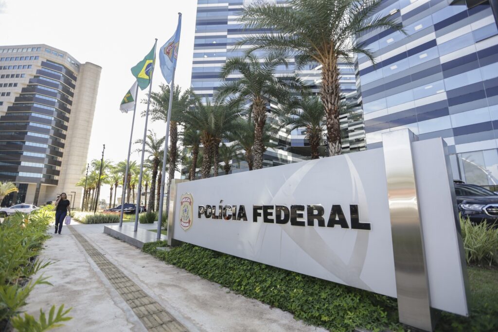 Entrada da sede da Polícia Federal com placa escrita "POLÍCIA FEDERAL", prédio moderno ao fundo, palmeiras e a bandeira do Brasil.