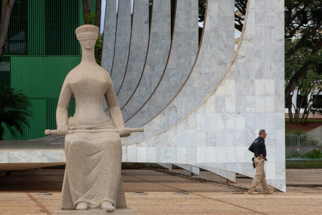 Estátua da Justiça com venda nos olhos e espada em frente a paredes curvas de mármore branco do Supremo Tribunal Federal, com um agente da "Polícia Judicial" caminhando ao fundo.