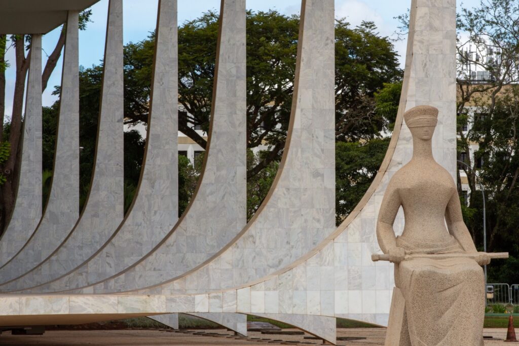 Estátua da Justiça, com vendas nos olhos e segurando uma espada, em frente às colunas curvas do Supremo Tribunal Federal.