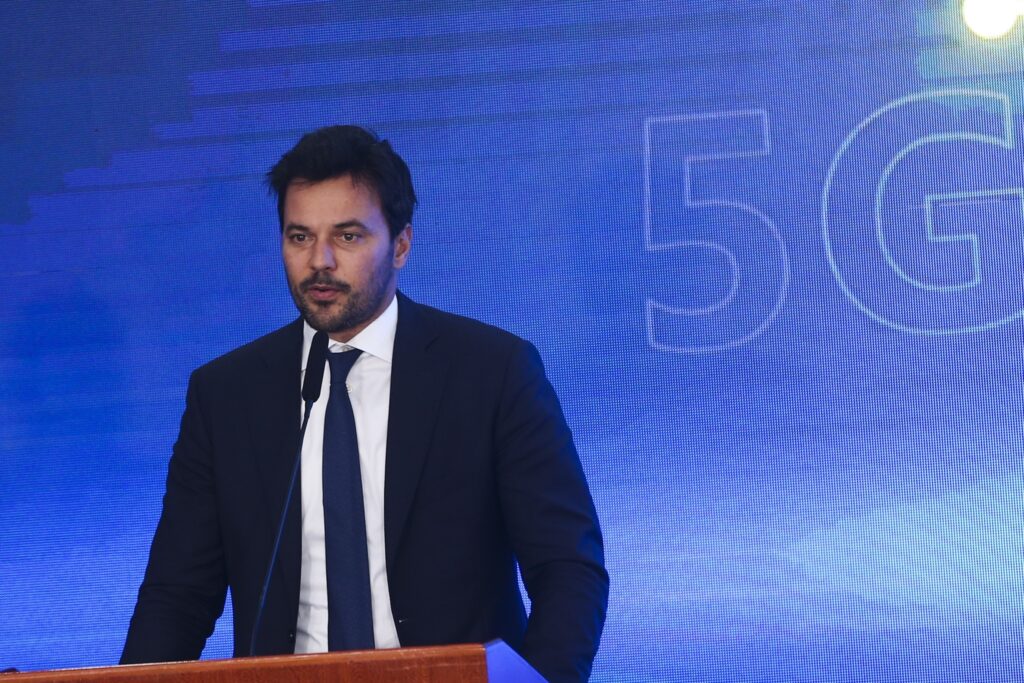 Homem de terno fala ao microfone em frente a um painel azul com a inscrição '5G'.