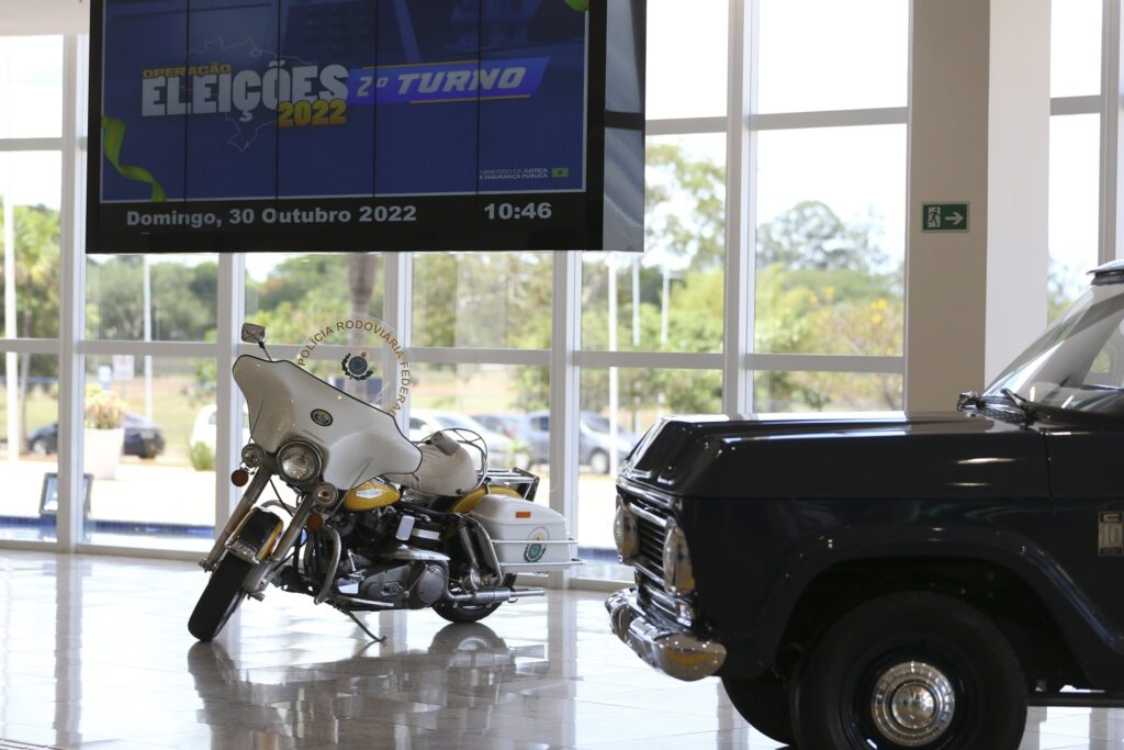 Motocicleta e veículo da Polícia Rodoviária Federal em ambiente interno, com painel eletrônico ao fundo exibindo "OPERAÇÃO ELEIÇÕES 2022 2º TURNO".