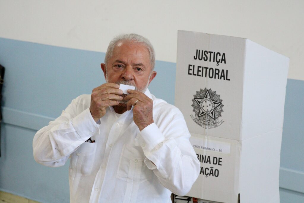 Luiz Inácio Lula da Silva segura um comprovante de votação ao lado de uma urna com os dizeres "JUSTIÇA ELEITORAL".