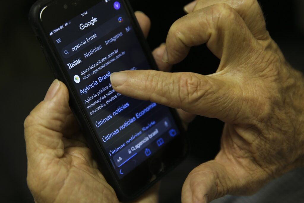 Mãos segurando um smartphone que exibe nos resultados de busca do Google o site da "Agência Brasil".