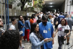 Pessoas aguardam em fila em uma seção eleitoral, com um funcionário vestindo uniforme azul com os dizeres "Justiça Eleitoral".