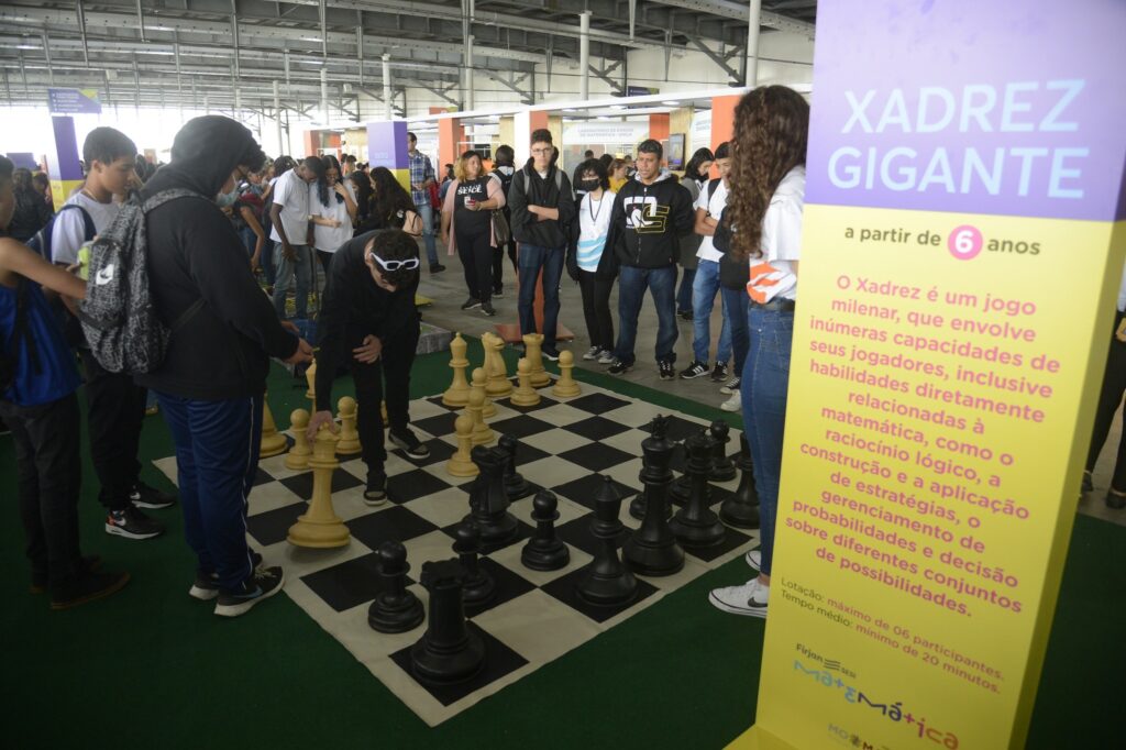 Jovens interagem com um tabuleiro de xadrez gigante ao lado de um banner com os dizeres "XADREZ GIGANTE" durante o 2º Festival Nacional da Matemática.