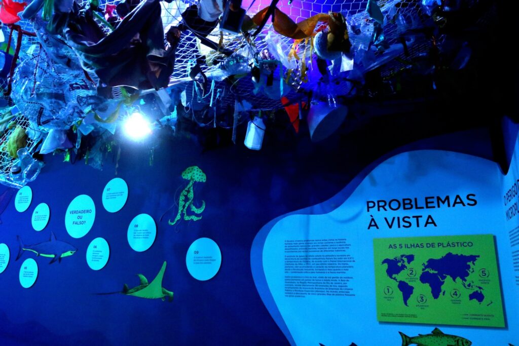 Exposição com rede de plásticos suspensa no teto e painéis educativos em parede azul com os títulos "PROBLEMAS À VISTA" e "AS 5 ILHAS DE PLÁSTICO".