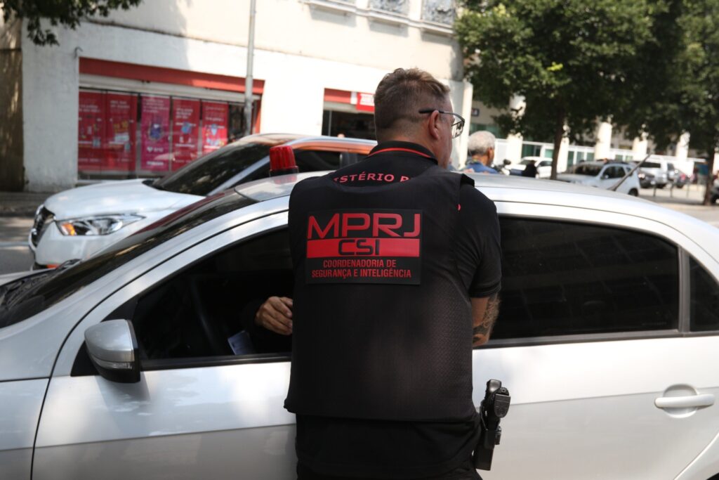 Agente do MPRJ de costas, usando colete com os dizeres "MPRJ CSI Coordenadoria de Segurança e Inteligência", ao lado de um carro branco em rua urbana.