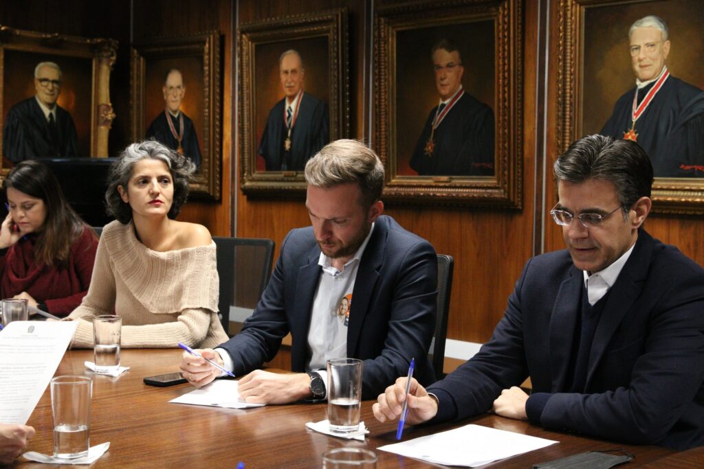 Quatro pessoas sentadas em uma mesa de reuniões em ambiente formal, com retratos de autoridades ao fundo e papéis para assinatura sobre a mesa.