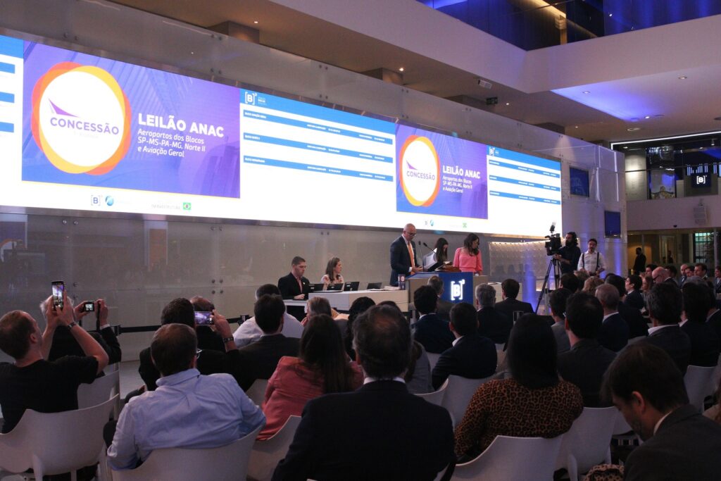 Pessoas assistem a um evento com painéis digitais com os dizeres "LEILÃO ANAC Aeroportos dos Blocos SP-MS-PA-MG, Norte II e Aviação Geral".