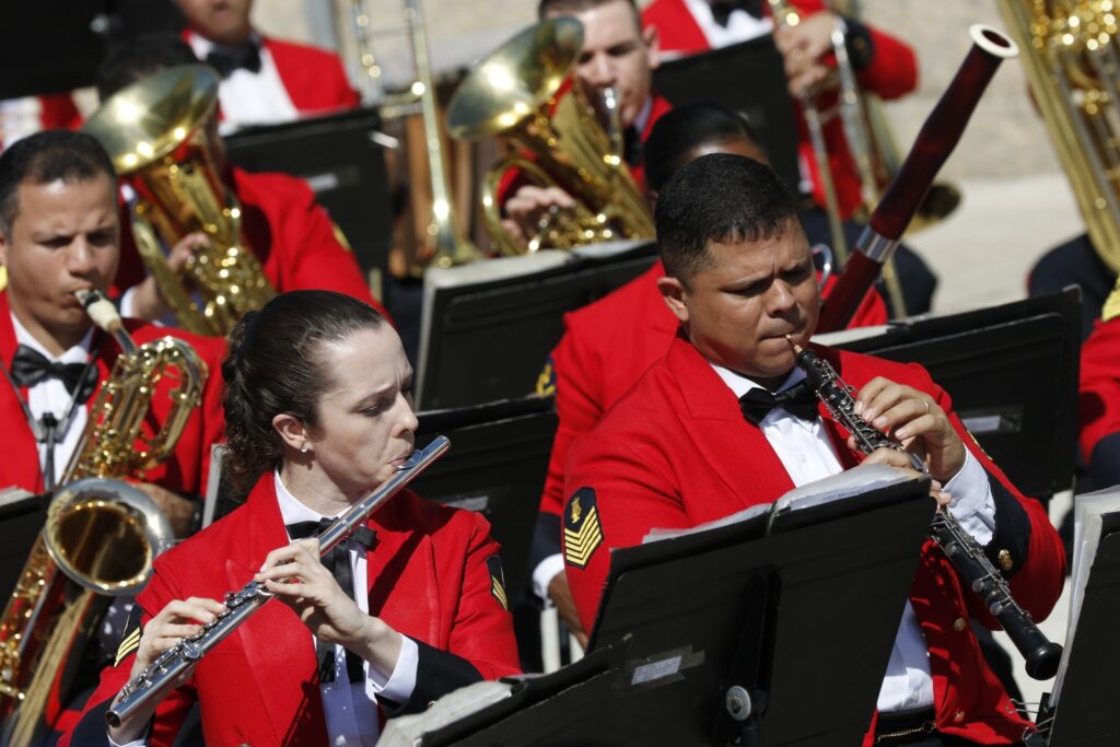 Músicos da Banda Sinfônica do Corpo de Fuzileiros Navais, vestindo uniformes vermelhos, tocam instrumentos de sopro ao ar livre.