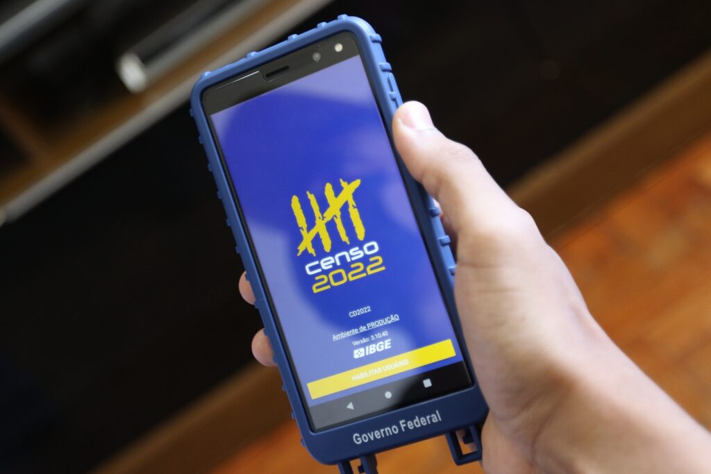 Mão segura celular com capa azul exibindo a tela do aplicativo "Censo 2022" do IBGE.