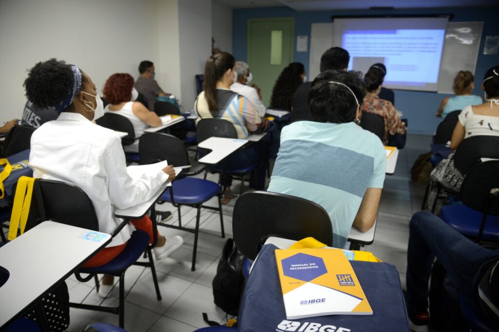 Pessoas sentadas em sala de aula assistem a treinamento, com um manual do "Censo 2022" do IBGE em destaque sobre uma mochila em primeiro plano.
