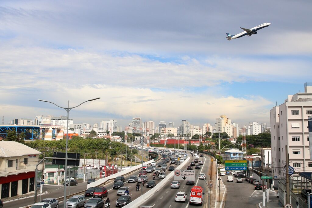 Avião da companhia Azul voa baixo sobre avenida movimentada com trânsito de carros, ambulância e placas indicando "Jabaquara" e "Imigrantes".