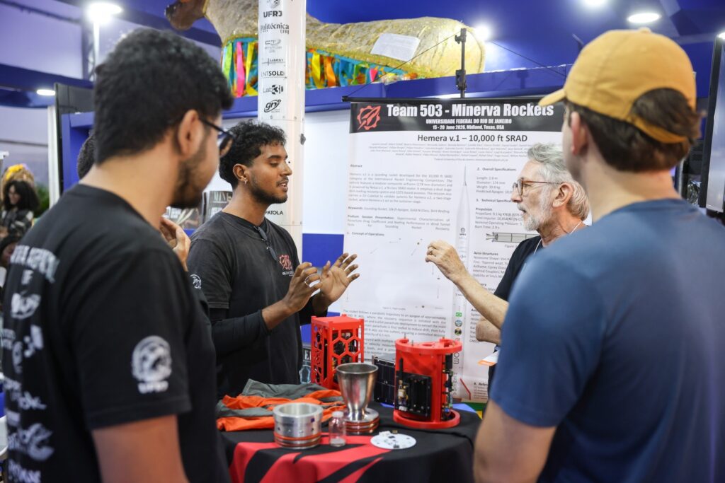 Homem gesticula ao explicar componentes de foguete para visitantes em um estande com o banner "Team 503 - Minerva Rockets" da Universidade Federal do Rio de Janeiro.