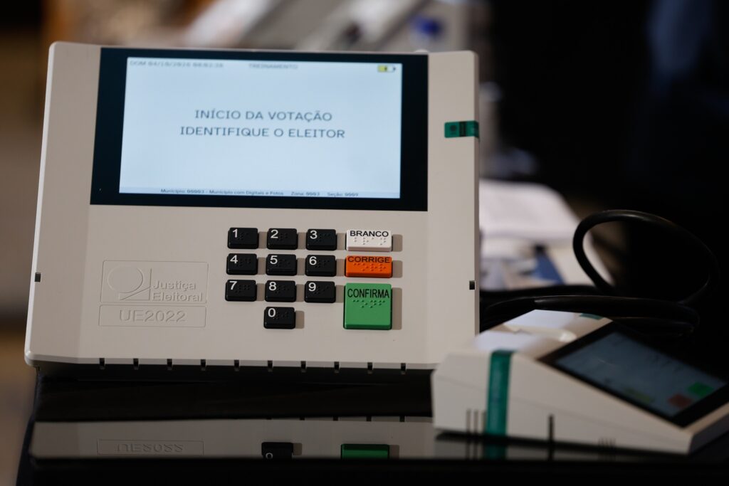Urna eletrônica com a mensagem "INÍCIO DA VOTAÇÃO IDENTIFIQUE O ELEITOR" na tela, ao lado de um leitor biométrico.
