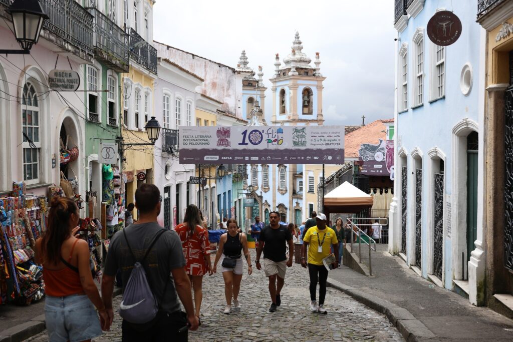 Pessoas caminham por rua de pedras no centro histórico de Salvador, sob faixa com os dizeres '10ª flipelô - Festa Literária Internacional do Pelourinho'.