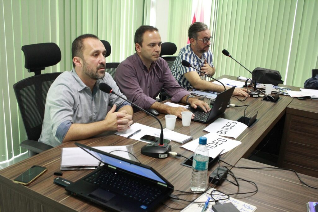 Três homens sentados em uma mesa de reunião com notebooks, microfones e papéis com a sigla 'BNDES'.