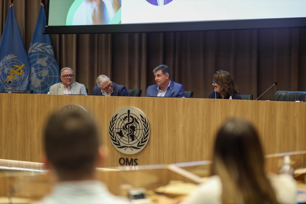 Quatro pessoas sentadas atrás de um pódio com o logotipo da "OMS" durante uma reunião em sala com bandeiras da ONU e da OMS.