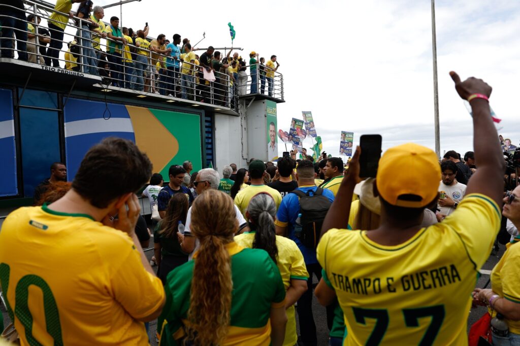 Multidão com camisas amarelas em comício político ao lado de um trio elétrico; um homem em destaque veste camiseta com os dizeres "TRAMPO E GUERRA 77".