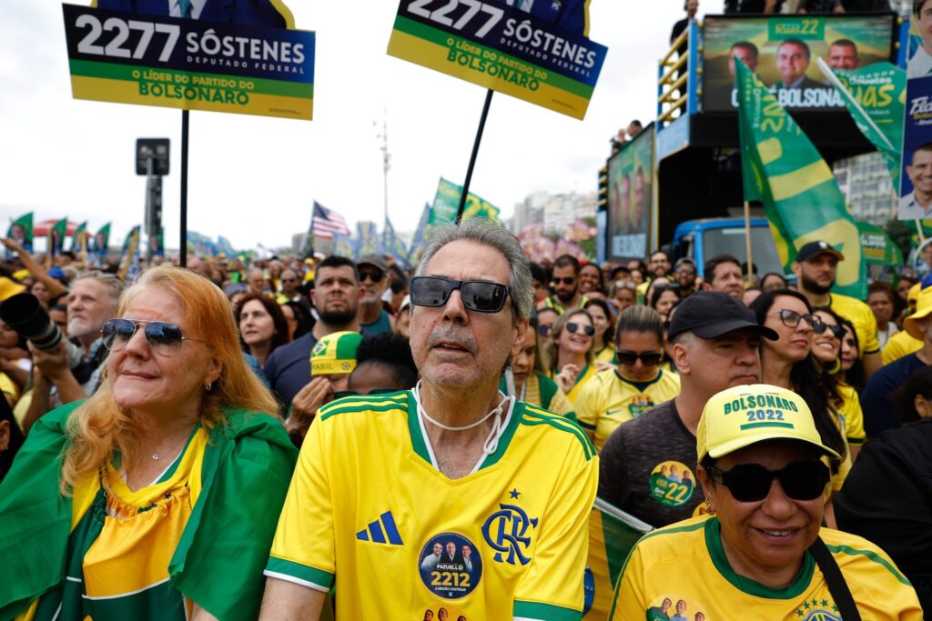 Multidão de apoiadores em comício político usando roupas nas cores do Brasil, com boné escrito "BOLSONARO 2022" e cartazes de "SÓSTENES DEPUTADO FEDERAL".