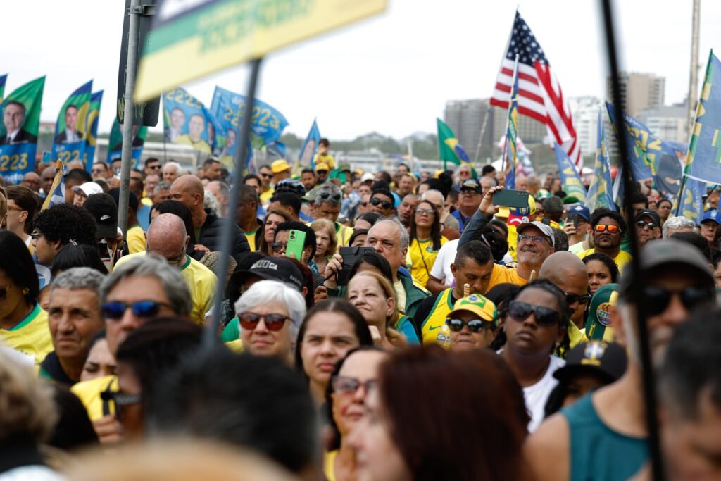 Multidão de apoiadores, muitos vestindo camisas amarelas, em comício político com bandeiras do Brasil, dos Estados Unidos e faixas com o número "22".