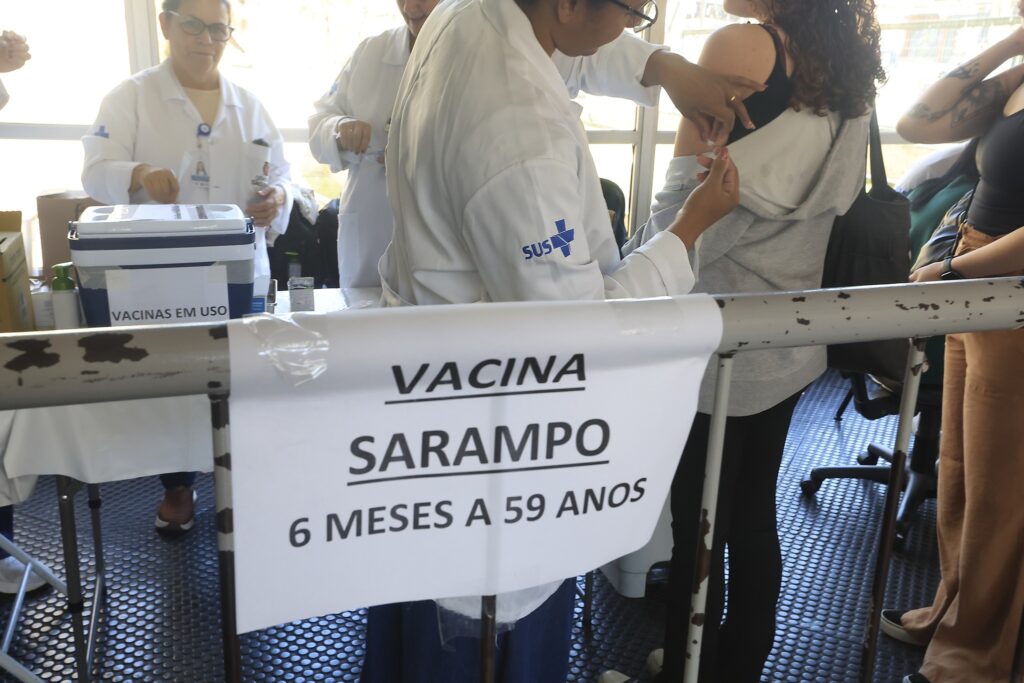 Profissional de saúde aplica vacina no braço de uma pessoa em um posto com a placa "VACINA SARAMPO 6 MESES A 59 ANOS".